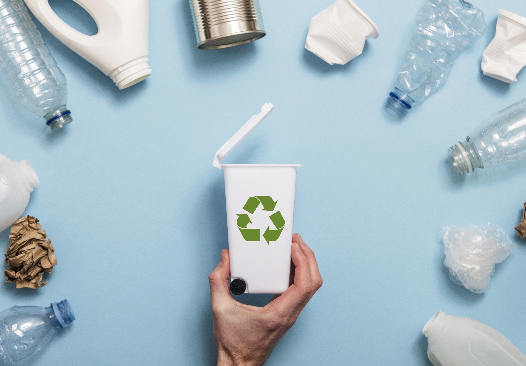 hand holding toy recycling bin surrounded by plastic jugs, plastic bottles, aluminum cans, paper, and more on blue background