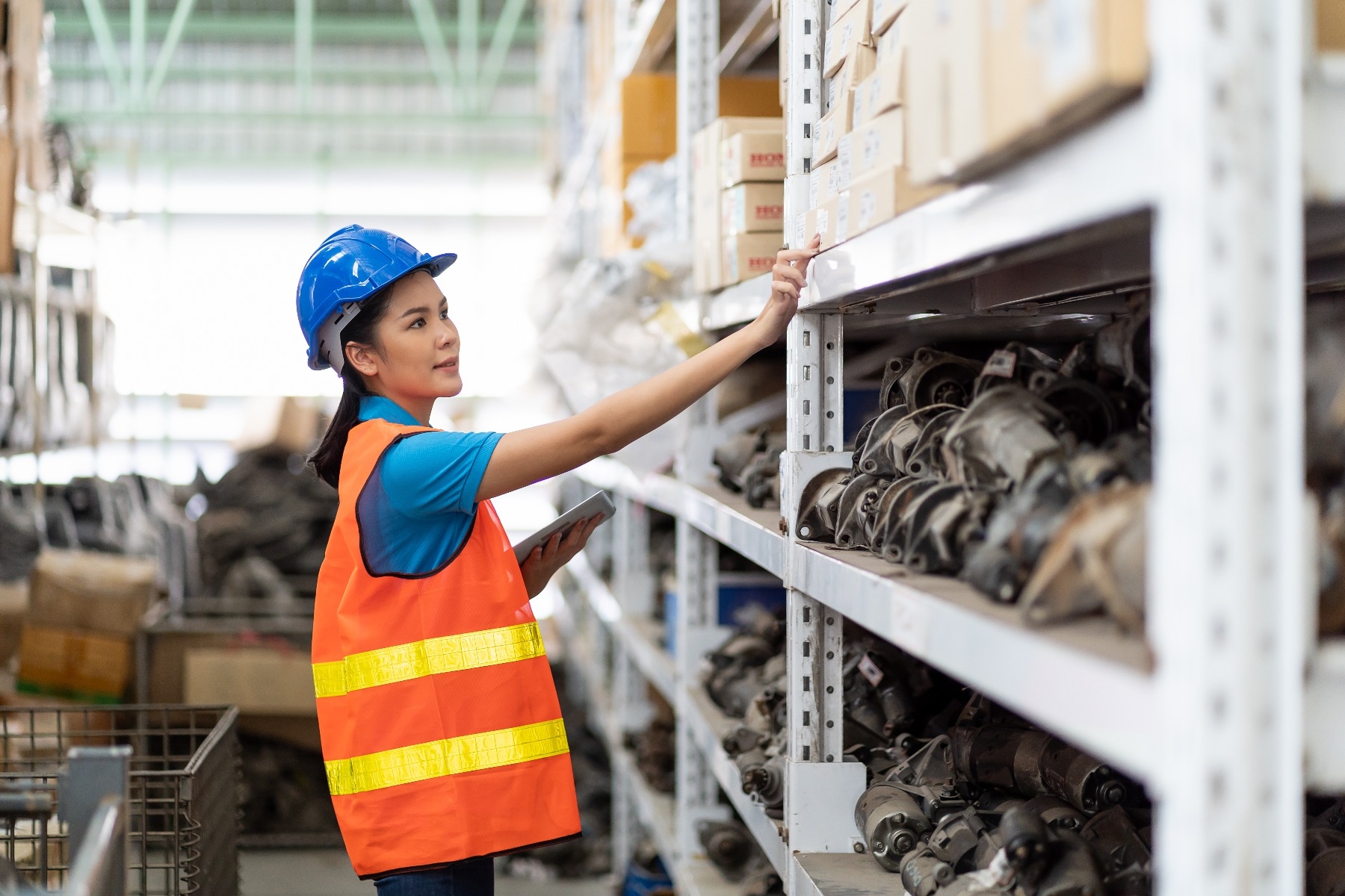 woman in safety vest and hard hat selecting car parts from warehouse shelves