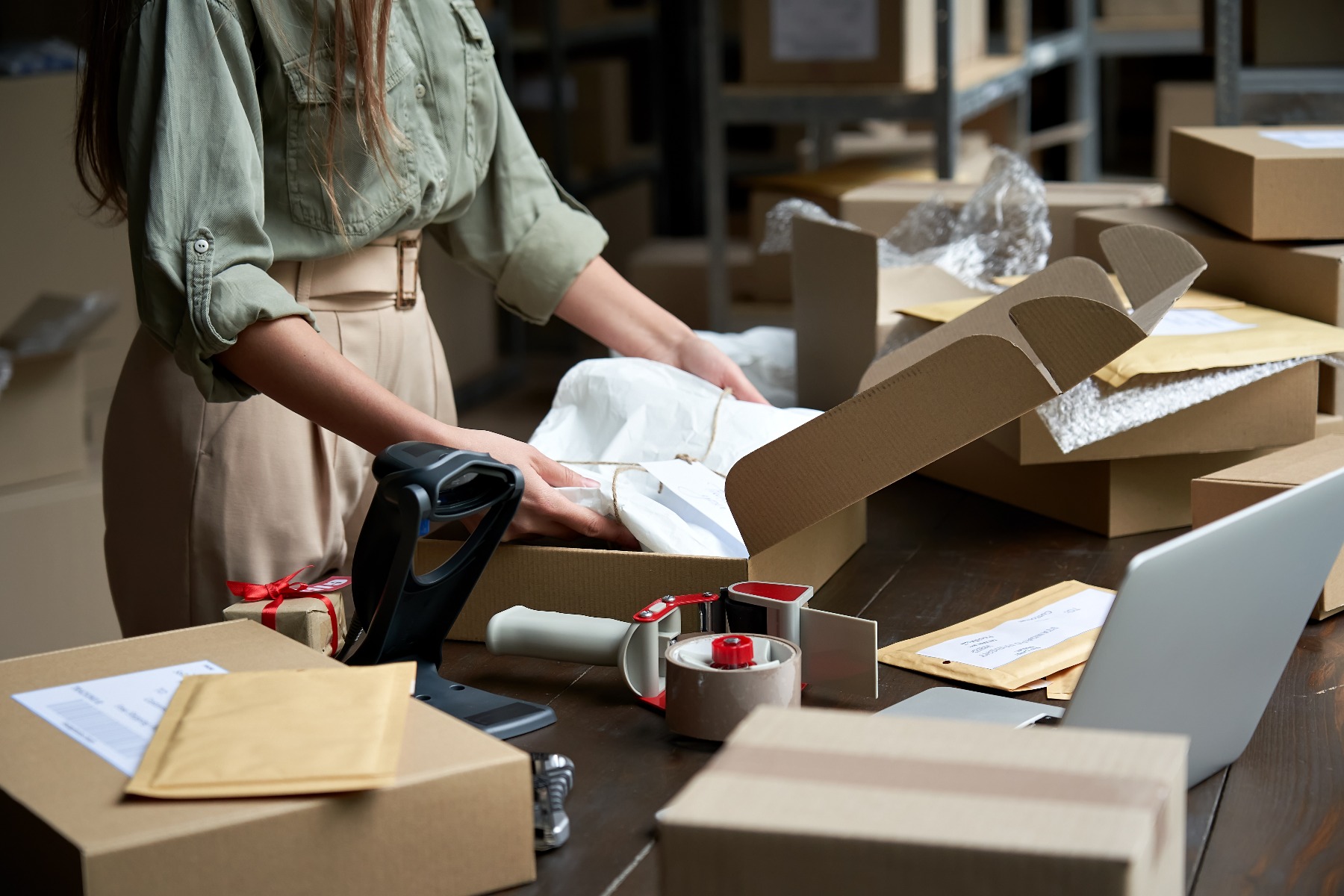 woman placing item wrapped in tissue paper into corrugated box with packaging supplies on table around it